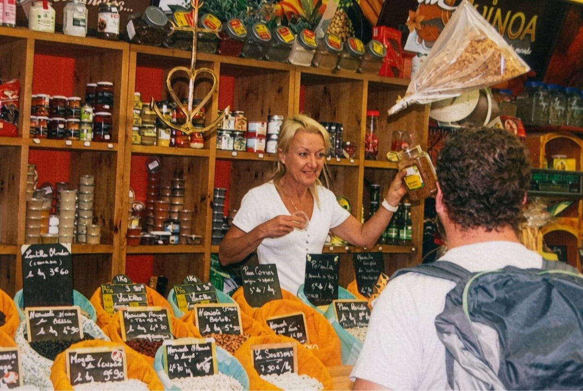 L'étal d'épices et légumineuses des Halles de Nîmes, berceau d'Épices Moralès — la fondatrice conseille un client parmi ses sacs de haricots, lentilles et aromates.