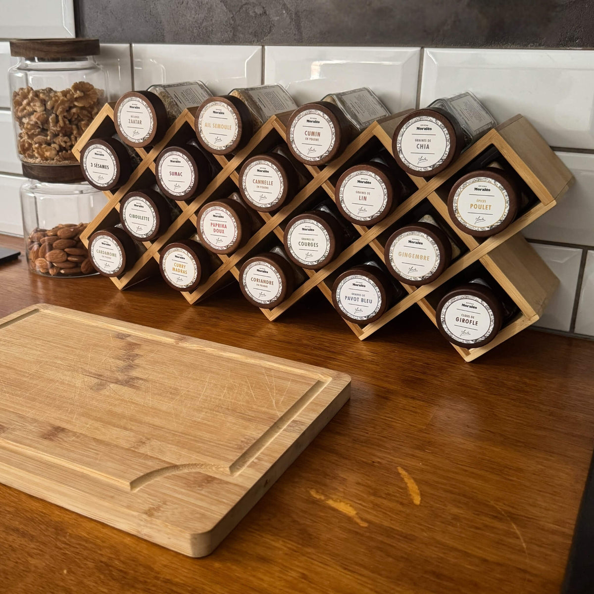Présentoir à épices en bois Épices Moralès garni de 18 fioles, posé en cuisine près d’une planche à découper.