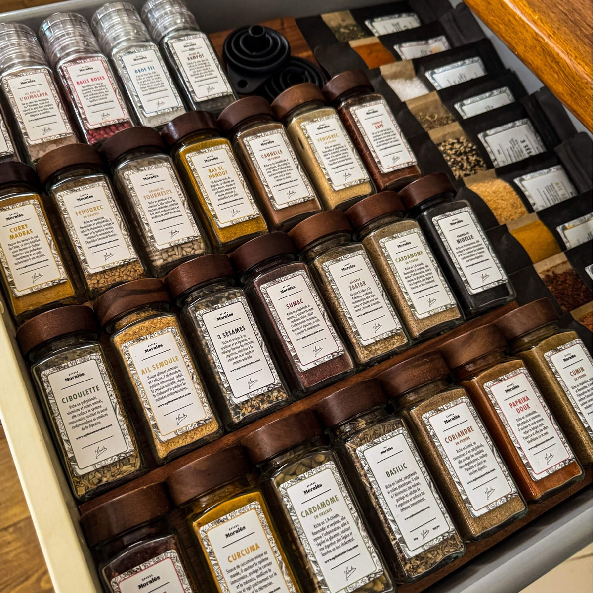 Tiroir de cuisine ouvert, fioles Épices Moralès étiquetées et rangées en épi, avec l’entonnoir pliable.